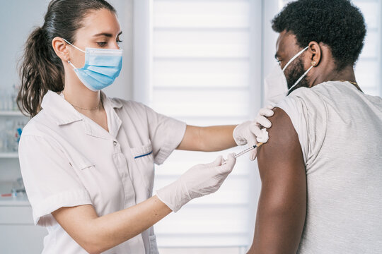 Female medical specialist in protective uniform, latex gloves and face mask vaccinating African American man patient in clinic during coronavirus outbreak