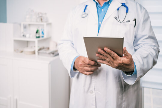 Unrecognizable cropped medic in robe using tablet in clinic