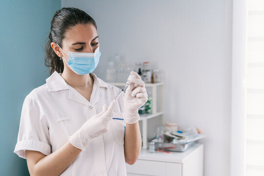 Female doctor in latex gloves and face shield filling in syringe from bottle with vaccine preparing to vaccinate patient in clinic during coronavirus outbreak