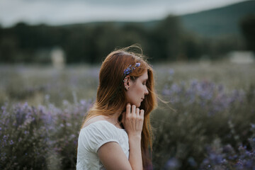 Side view of gentle female with flowers in hair sitting in blooming lavender field and enjoying nature with closed eyes