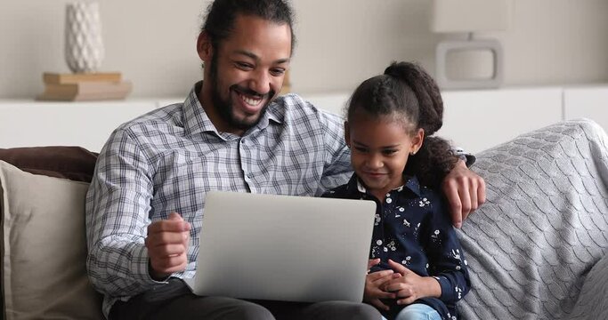 Young African father and little 5s girl using laptop seated on sofa spend time at home with wireless tech, watch cartoons, caring parent teach daughter to use modern device. Weekend leisure concept
