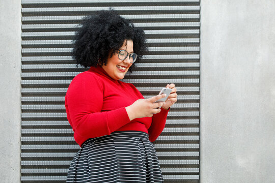 Side view of plump smiling female in bright wear text messaging on cellphone while leaning on ribbed wall in town