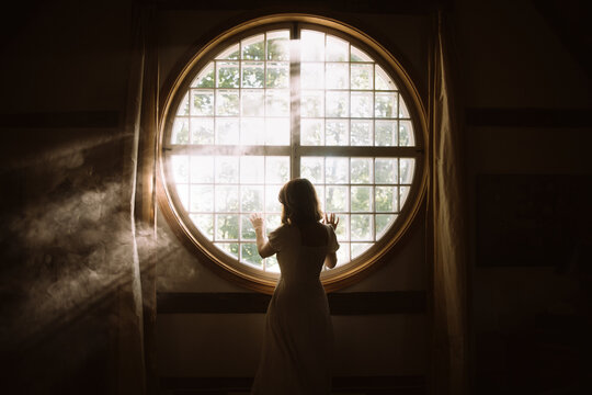 Fototapeta Back view of unrecognizable gentle female touching fence on round shaped window in house in sunlight