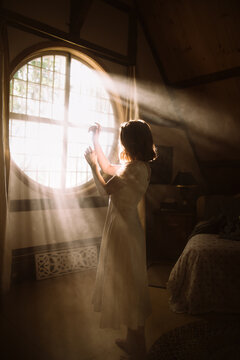 Side view of unrecognizable female in dress standing on couch against round shaped window in house on sunny day