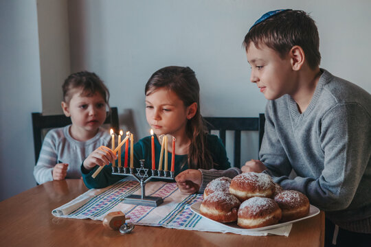 Brother and sisters siblings lighting candles on menorah for Jewish Hanukkah holiday at home. Children celebrating Chanukah festival of lights. Dreidel and Sufganiyot donuts in plate on table.
