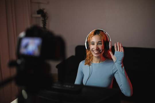 Smiling female gamer in headphones waving hand while recording video on professional camera for social media blog