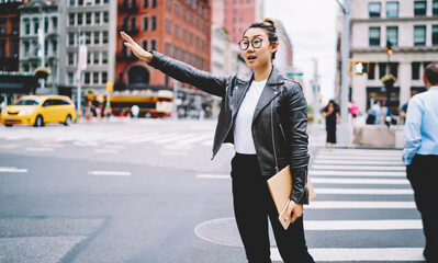 Chinese woman with education sketchbook gesturing for catch cab transport and get to destination in urban city, trendy dressed hipster girl in optical spectacles waving at street with traffic