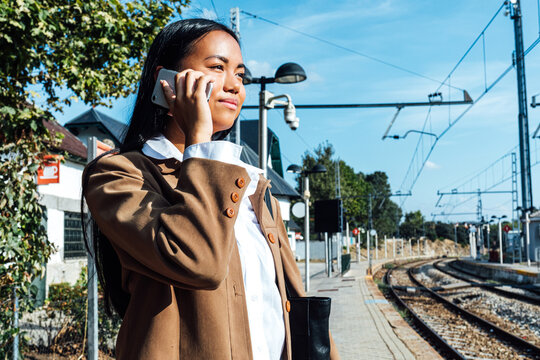 From below side view of content ethnic female traveler standing on platform at railroad station and talking on smartphone