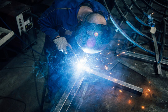 Crop faceless worker in gloves and uniform welding metal details on table near constructions in factory