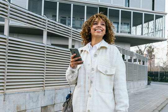 Optimistic African American female with Afro hairstyle browsing on smartphone while standing against metal wall in urban area in city