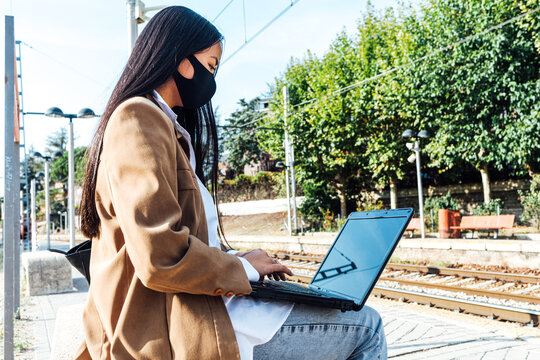 Side view of Asian female in mask sitting on bench at railway station and browsing laptop while waiting for train