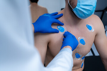 Crop medic applying electrodes on chest of unrecognizable male patient in mask for ECG test