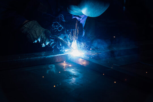 Crop faceless worker in gloves and uniform welding metal details on table near constructions in factory