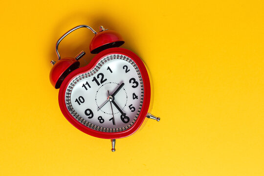 Red metal alarm clock in shape of heart placed on vibrant yellow background in studio