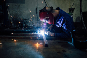 Crop faceless worker in gloves and uniform welding metal details on table near constructions in factory