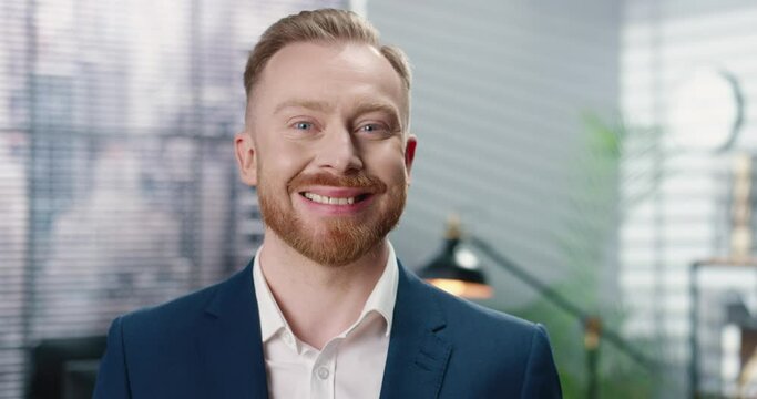 Close up portrait of joyful handsome young red-haired businessman with beard looking at camera and smiling in cabinet in good mood. Cheerful male employee with smile on face at work. Business concept