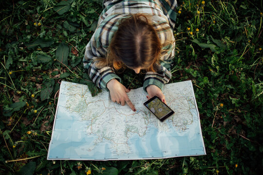 From above back view of unrecognizable female tourist with cellphone watching route map while lying on grass in countryside