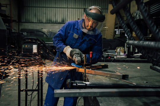 Workman in protective mask and blue workwear grinding metal part with angle grinder at workbench in professional garage