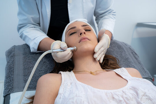 Cropped unrecognizable professional cosmetologist using special equipment and doing microdermabrasion facial treatment for female client in modern beauty clinic