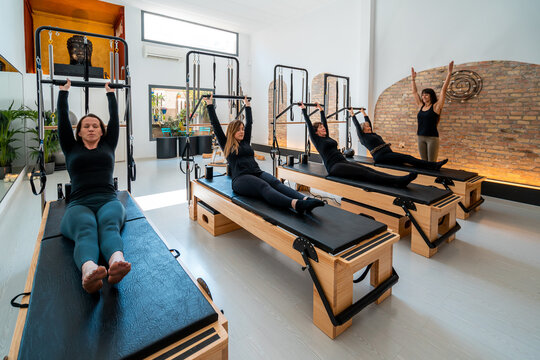 Company of females in sportswear stretching body on pilates reformers during training under supervision of professional instructor