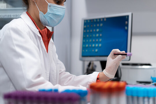 Side view of unrecognizable female medic in mask and gloves holding tube with blood sample in modern lab in clinic