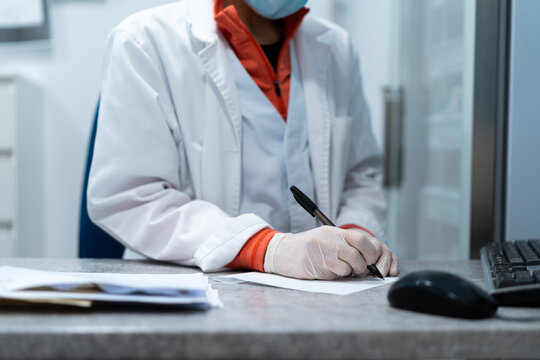 Cropped unrecognizable female doctor in mask and uniform sitting at table in medical room and writing prescription on paper