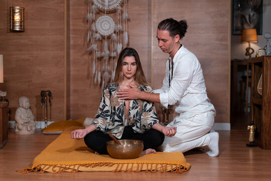 Zen teacher in white apparel touching chest of female with crossed legs during yoga practice near bowl gong