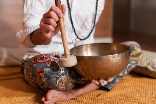 Crop unrecognizable Zen teacher with mallet playing Tibetan singing bowl while sitting with crossed legs on plaid