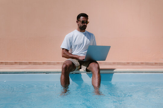 Male freelancer in sunglasses sitting at poolside and browsing netbook while working remotely on project during summer vacation