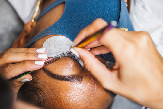 High angle of crop unrecognizable cosmetologist with tweezers applying fake eyelashes for extension on eye of ethnic client with face protective mask in salon during coronavirus pandemic