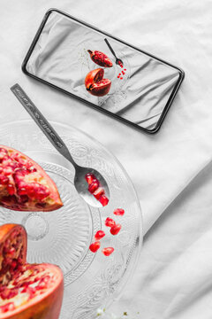 Top view of bright pomegranate seeds with spoon on stand near cellphone with photo on screen on white background