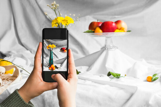 Crop anonymous person taking photo of fresh kumquats and blooming flower bouquet in vase on cellphone on white background