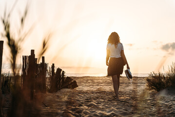 Back view of unrecognizable female in summer clothes walking towards sea along sandy beach on background of sundown sky