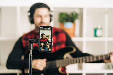 Tripod with cellphone screen representing photography of male musician in headset playing bass guitar in house