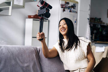 Smiling ethnic female vlogger recording video on photo camera while sitting on couch in living room