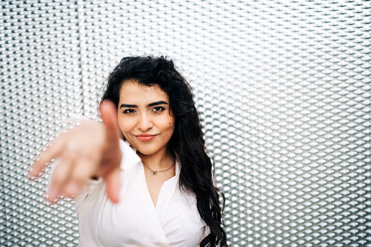 High angle of lovely curvy female with long hair pointing at camera while standing against metal mesh