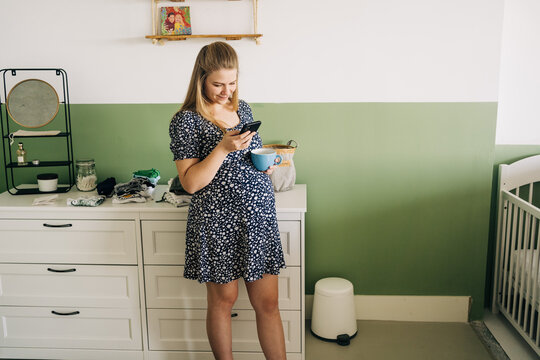 Young expectant female in ornamental wear with cup of hot drink chatting on cellphone in house