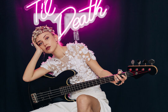 Energetic rebellious young woman in elegant white bridal dress and wreath with guitar in hand making horn gesture in studio with neon inscription