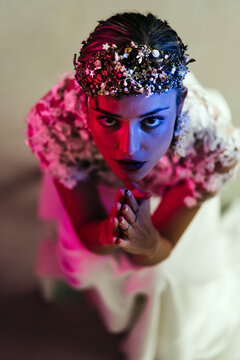 From above of elegant young female in white wedding gown with jewelry wreath looking at camera with praying hands in studio with neon illumination