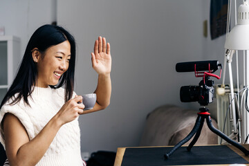 Smiling ethnic female blogger with cup of hot drink showing hello gesture while recording video on photo camera at home