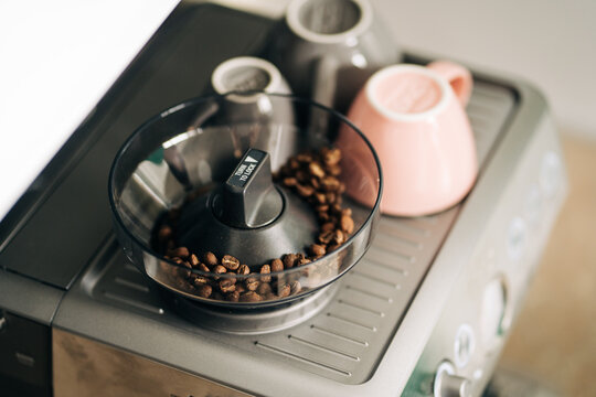 From above of roasted coffee beans in electric grinder against ceramic cups on rack of modern espresso maker at home