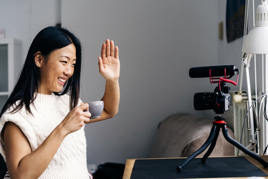Smiling ethnic female blogger with cup of hot drink showing hello gesture while recording video on photo camera at home