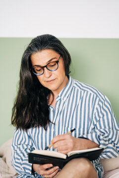 Middle aged cheerful female in striped shirt smiling and writing notebook on bed with smartphone