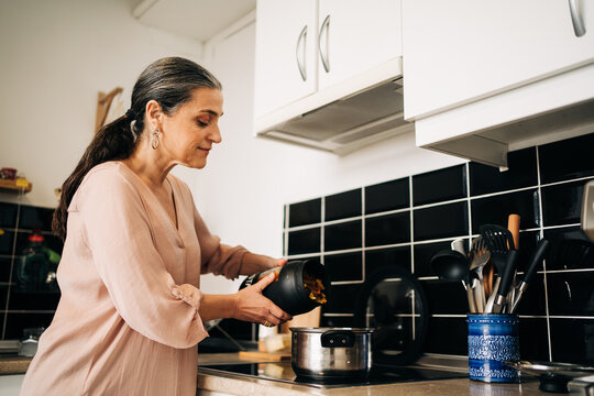 Side view of focused middle aged female adding fresh cut vegetables from container into pot while standing near stove in kitchen