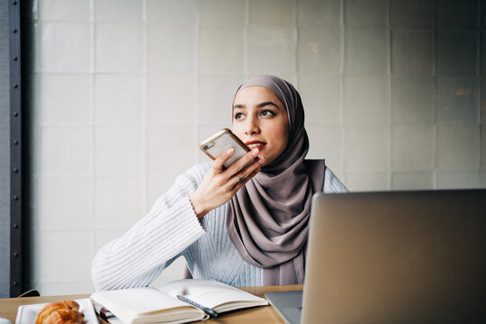 Content ethnic female freelancer in hijab recording audio message on smartphone while sitting at table in cafe and working remotely
