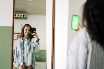 Cheerful middle aged female wearing striped blouse reflecting in mirror while taking self portrait on smartphone in room at home