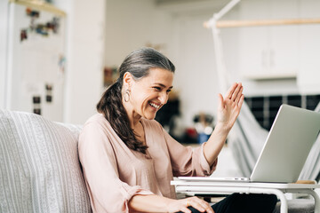 Positive middle aged female sitting on couch waving while having video chat on netbook in modern apartment