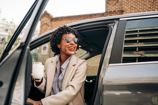 Happy African American female in fashionable sunglasses with thermal mug smiling and leaving silver prestige automobile