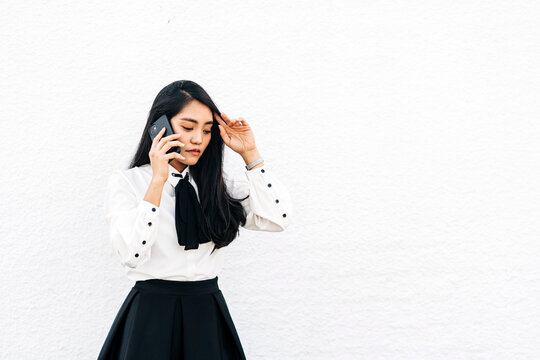 Teen ethnic Asian female in formal outfit standing near white wall speaking on the mobile phone