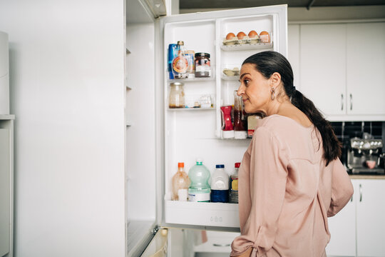 Side view of calm female with ponytail opening door of refrigerator with various products while standing in kitchen with white cupboards